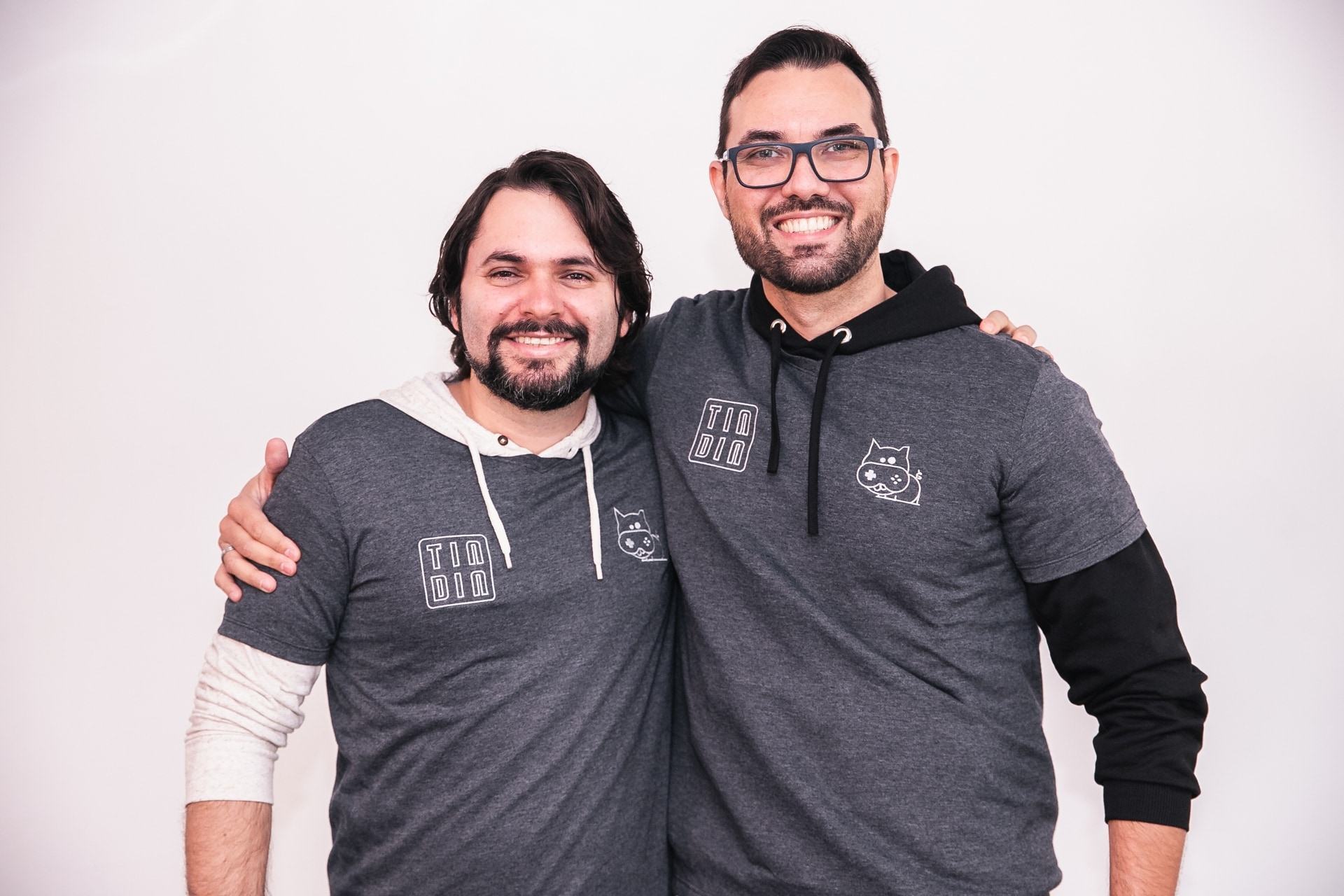 Dois homens brancos, de barba, abraçados. O da esquerda tem cabelo maior, liso; o da direita usa óculos. Ambos sorriem.