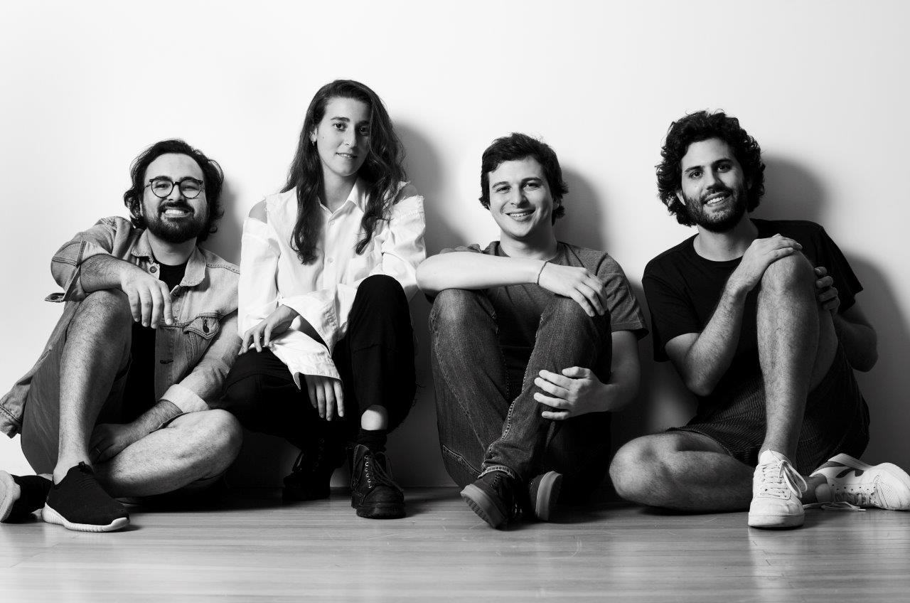 Quatro pessoas, três homens e uma mulher, sentados contra a parede enquanto olham para a câmera. Foto em preto e branco.