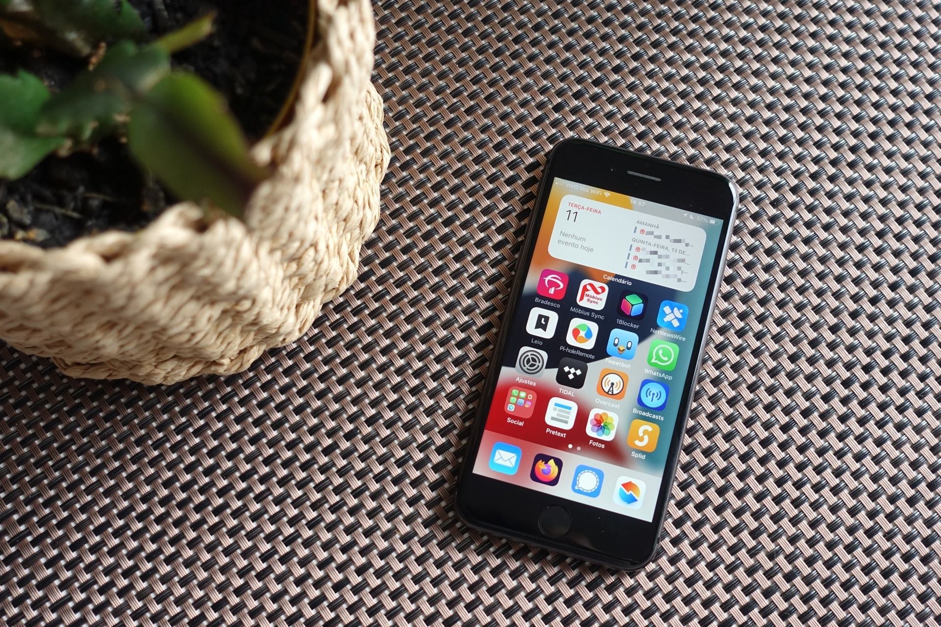 Foto de cima de um iPhone 8, com a tela ligada, sobre uma superfície marrom entrelaçada. À esquerda, uma planta com um vaso com cachepó de cipó.