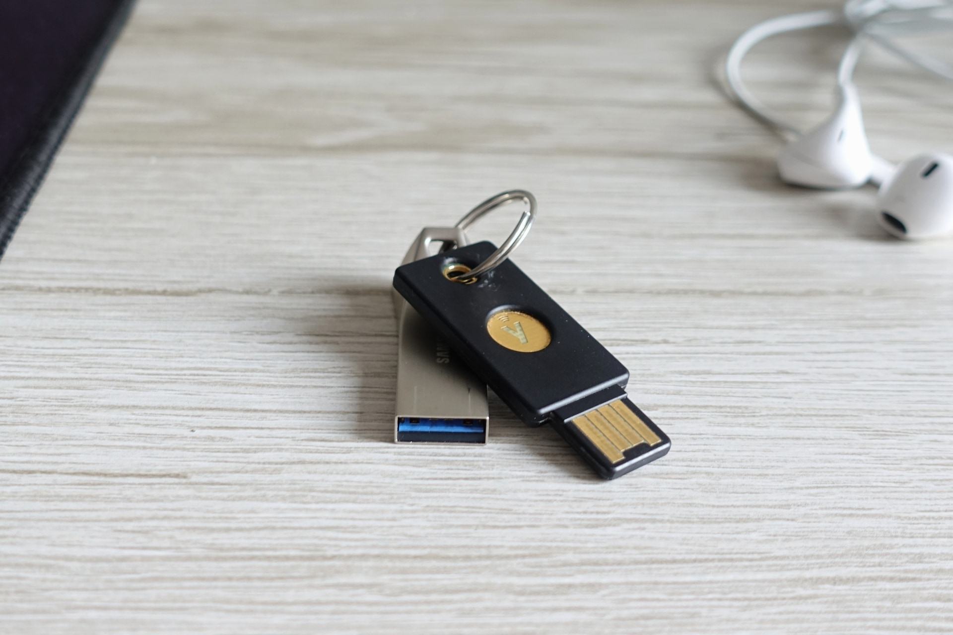 Foto em close de uma Yubikey preta e um pen drive prata unidos por uma argola/chaveiro. Ao fundo, desfocado, os fones dos EarPods.