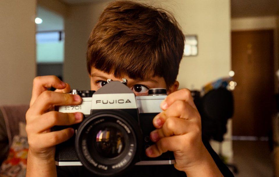 Menino segurando uma câmera antiga, analógica, com parte do rosto coberto — apenas olhos e topo da cabeça à mostra.