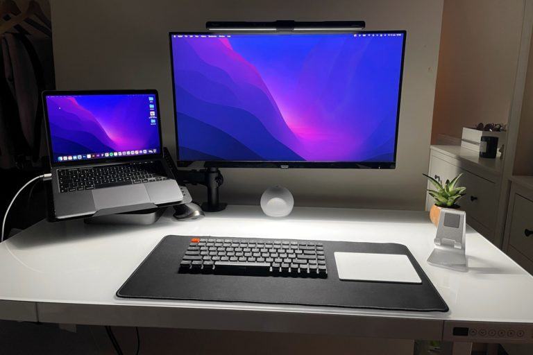 Mesa de trabalho com iluminação baixa, com monitor grande ao centro, notebook à esquerda e suporte para celular e planta pequena à direita. No centro, mousepad grande com teclado e trackpad da Apple.