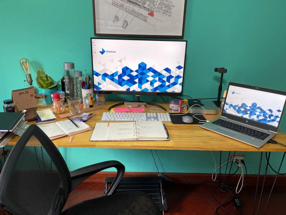 Visão frontal da mesa do Jorge, com o monitor e o notebook ligados, exibindo um papel de parede da Edelman.