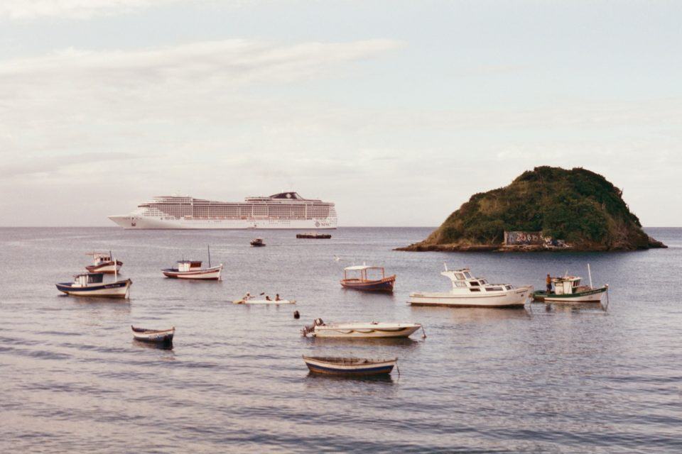 Vários barquinhos no mar. Ao fundo, um navio de cruzeiro e uma pequena ilha. Foto com efeito antigo (tirada com câmera analógica).