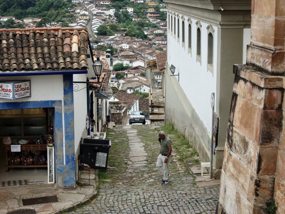 Foto de uma ladeira, de cima, com muitas casinhas ao fundo. Em primeiro plano, um senhor com a máscara pendurada na orelha e uma portinha vendendo pedras preciosas.