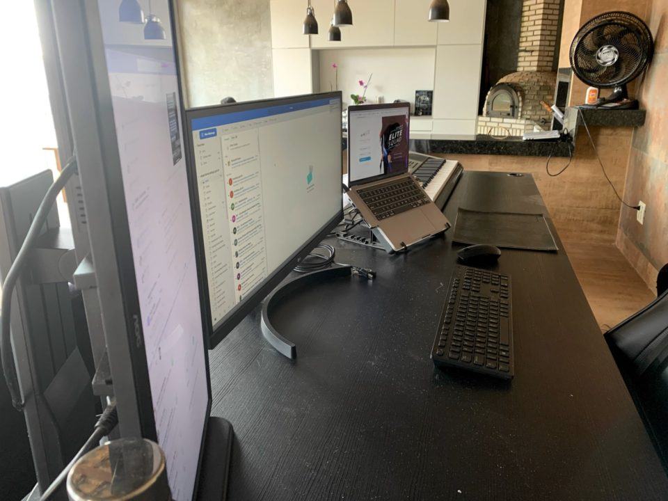 Vista do outro lado (dos monitores) do escritório do Rafael. Ao fundo, uma parede branca decorada e uma churrasqueira.