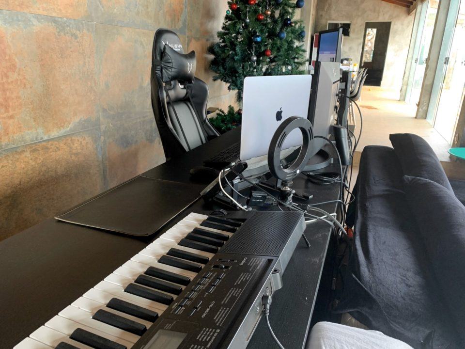 Vista de trás da mesa do Rafael, mostrando um teclado, o MacBook Pro dele, uma cadeira gamer preta e uma árvore de Natal.