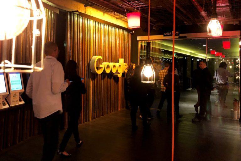 Foto de um hall, pouco iluminado, do escritório do Google em São Paulo, com o logo da empresa na parede e pessoas andando em volta.