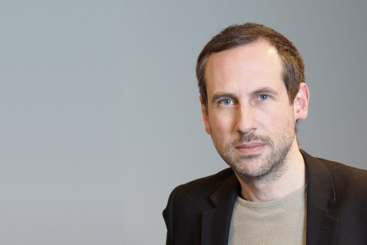 Homem branco, de cabelo castanha curto e barba por fazer, com camiseta bege e casaco preto, olhando para a câmera. Fundo em azul/marrom claro.