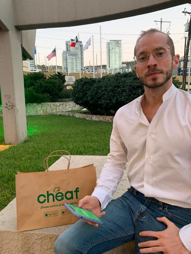Homem branco, de óculos redondos e barba por fazer, vestindo jeans e camisa branca, segurando um celular. A seu lado, uma sacola de papel da Cheaf.