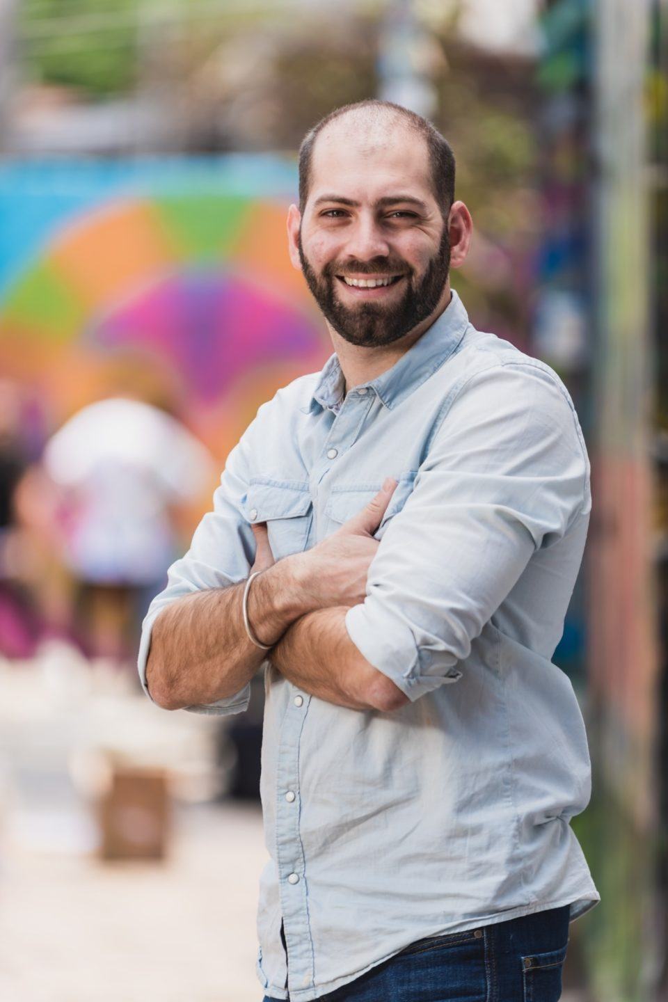 Homem branco, careca, de barba e camisa azul claro, com os braços cruzados, sorrindo e olhando para a câmera.