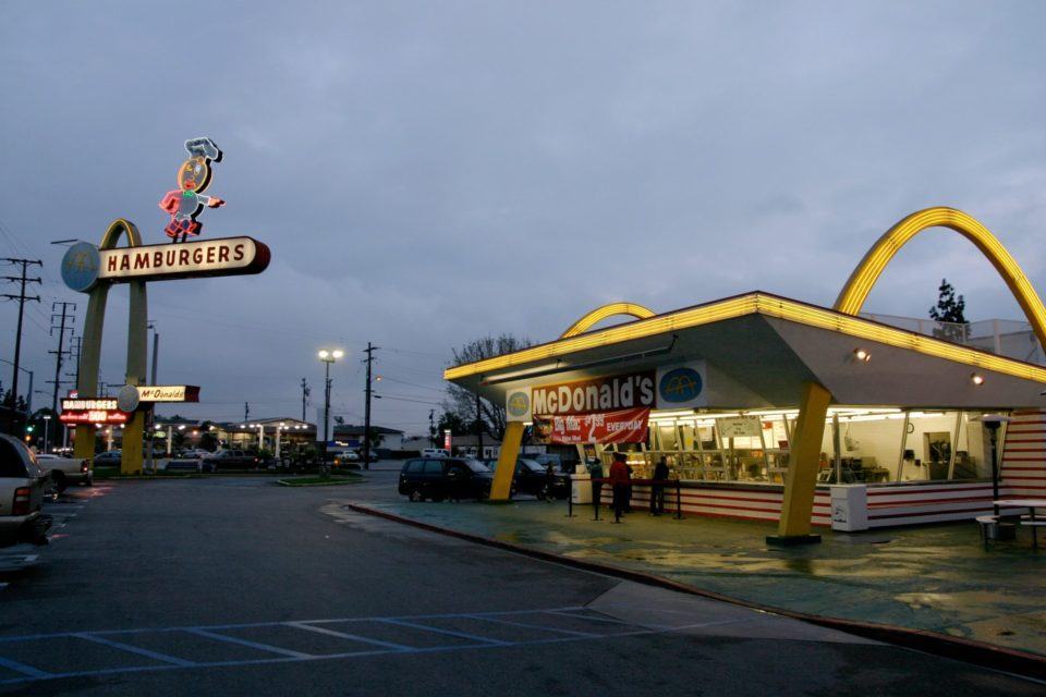 Fachada de um restaurante do McDonald's, mostrando parte do estacionamento e uma placa grande com um chef apontando e a palavra “hambúrguer” embaixo.