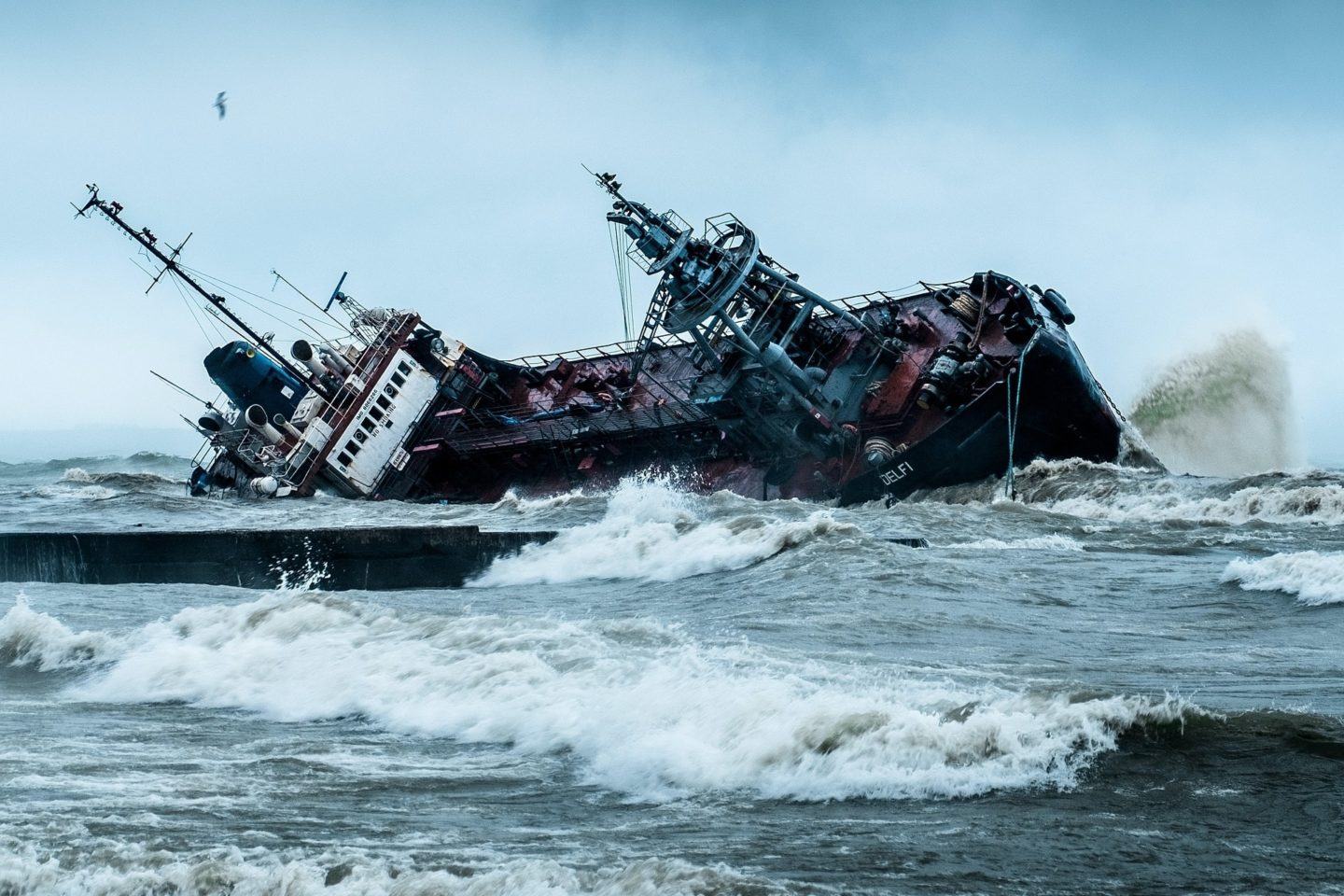 Navio tombado, com ondas em primeiro plano e um céu nublado ao fundo.