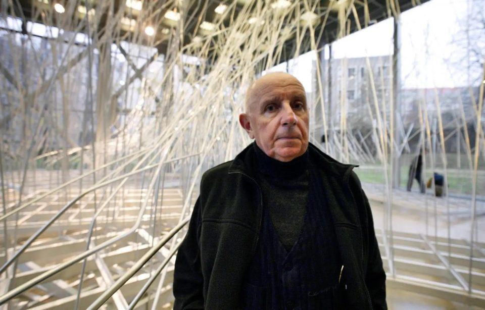 Homem branco, careca, com roupa pesada preta, em frente a uma instalação artística cheia de cabos e tubos brancos.