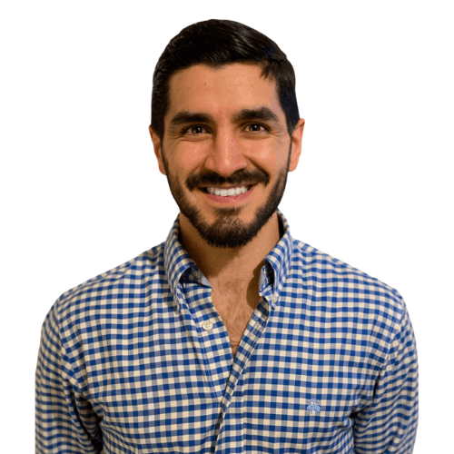 Homem branco, de cabelo curto e barba pretos, vestindo camisa xadrez azul e branca, sorrindo e olhando para a câmera.