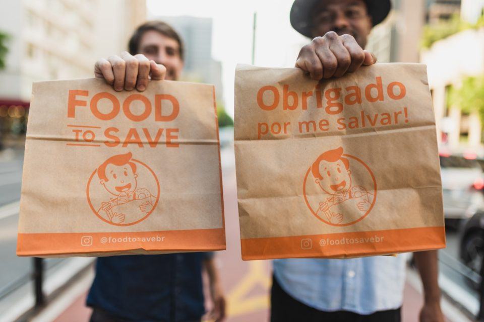 Dois homens, fora de foco, segurando sacolas de papel da Food to Save, focadas em primeiro plano.