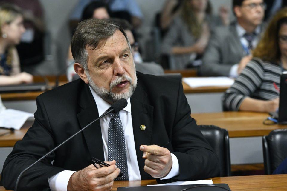 Homem branco, de barba e cabelo grisalhos, vestindo terno, sentado atrás de uma bancada e falando ao microfone.