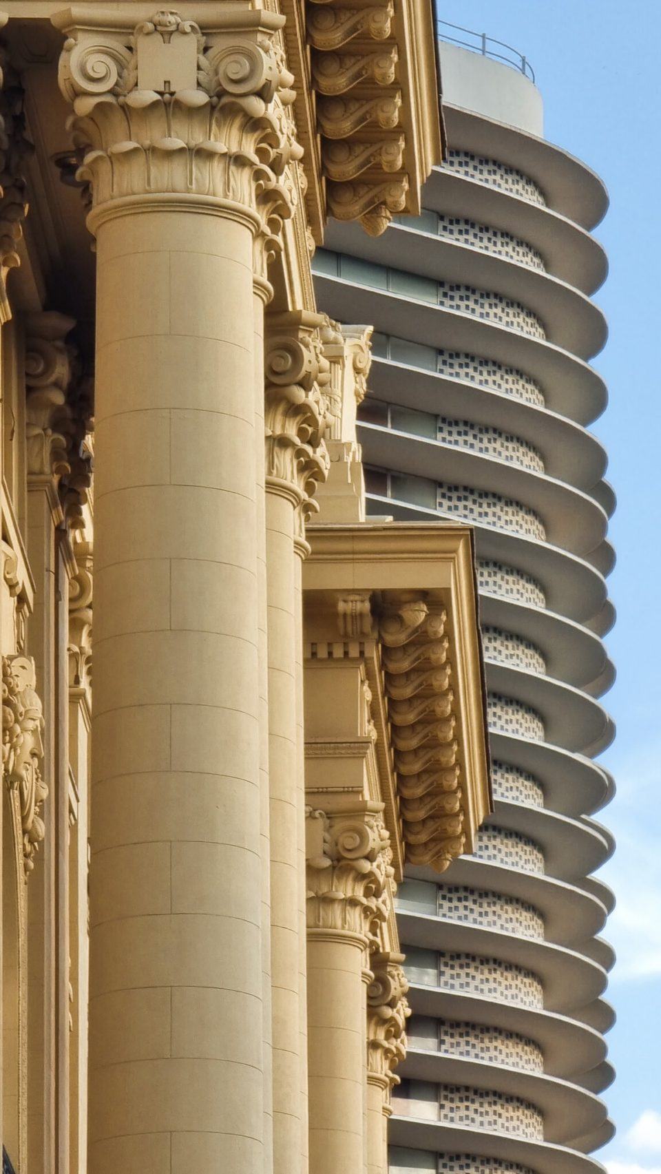 Duas fachadas de prédios, um clássico, outro mais moderno, no detalhe, contra o céu azul.