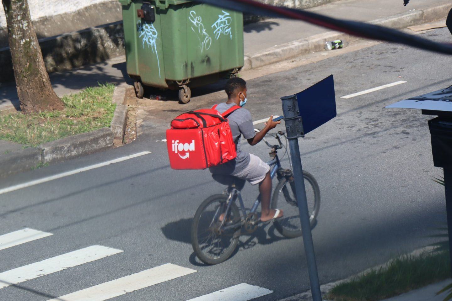 Homem negro andando de bicicleta, com uma mochila do iFood nas costas, manuseando um celular, máscara no queixo. Ao fundo, na rua, uma lixeira pixada.