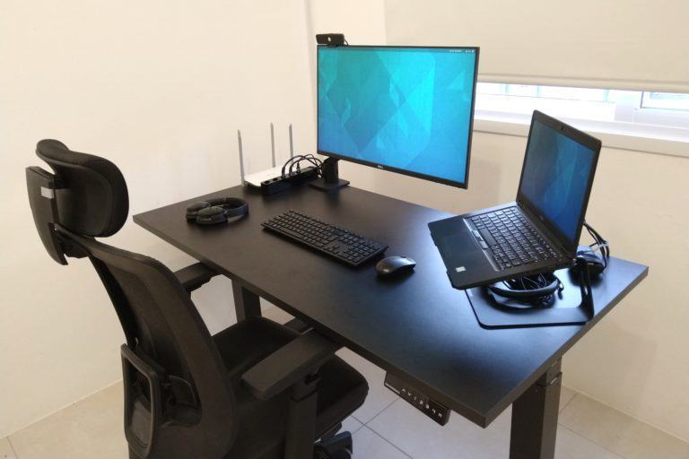 Foto na diagonal, de cima, de uma mesa preta, com notebook, monitor e acessórios, e uma cadeira grande preta, em um ambiente claro.