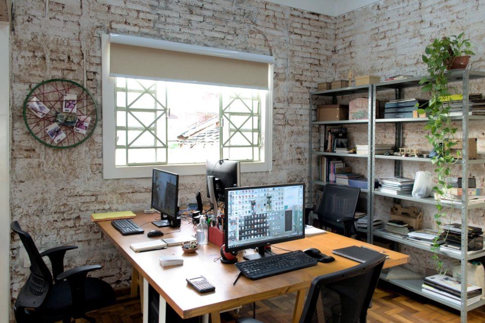 Escritório visto por outro ângulo, da ponta da mesa, com uma janela aberta ao fundo e uma estante decorada à direita.