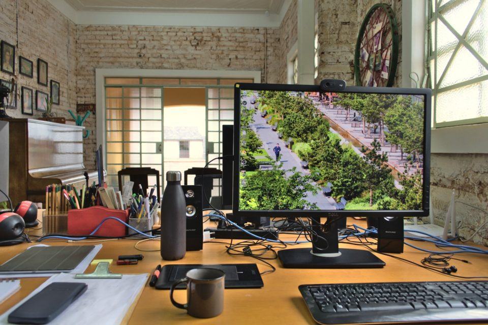 Foto em close de uma das estações de trabalho, mostrando o monitor, teclado, xícara e alguns outros objetos sobre a mesa. Ao fundo, a porta de vidro aberta e um piano fechado à esquerda.