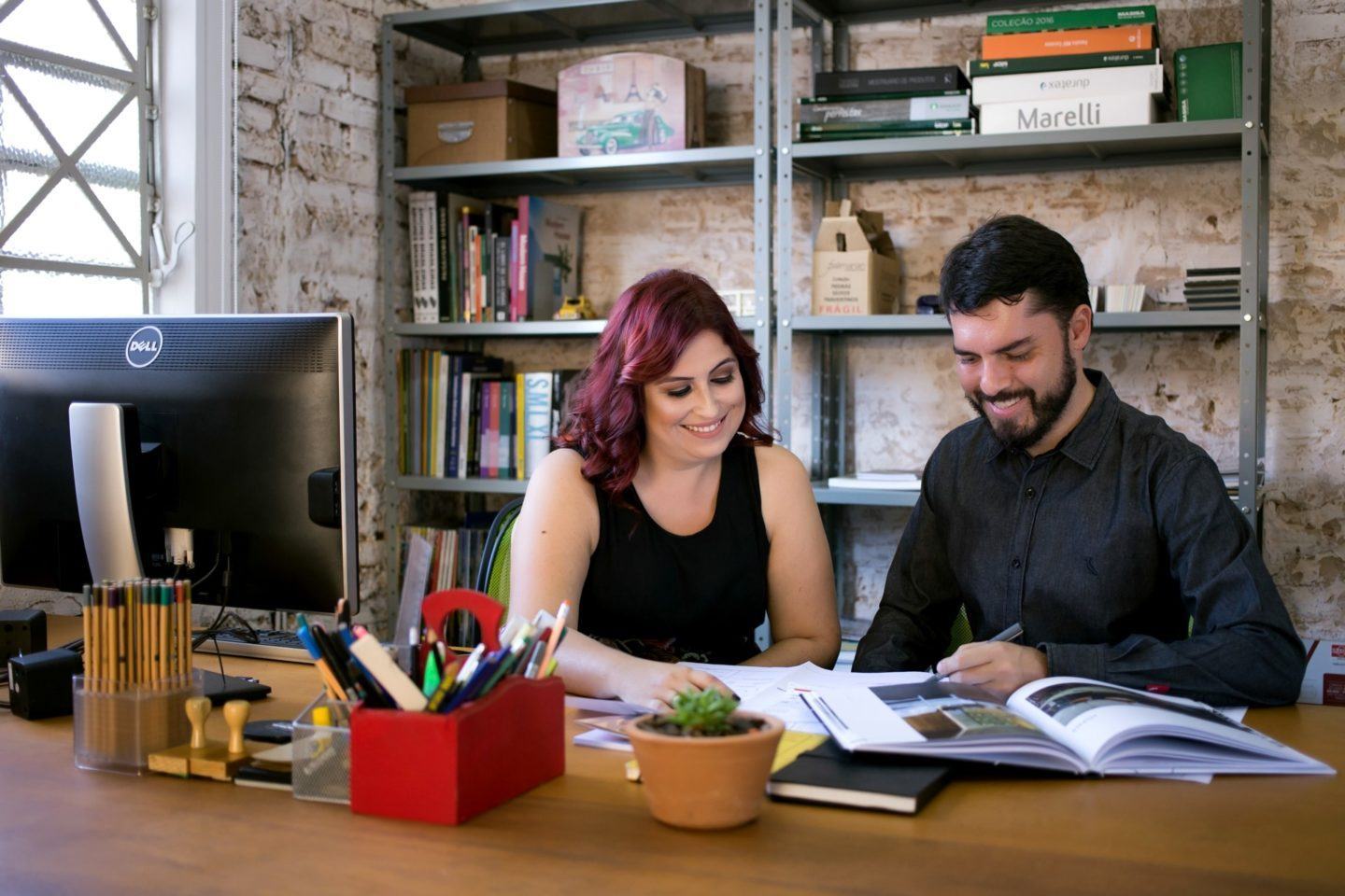 Mulher branca, de cabelo comprido, ondulado e roxo, com um vestido preto; e um homem branco, de cabelo e barba pretos, com camisa preta. Ambos sentados, olhando para papéis sobre uma mesa. À esquerda, um monitor de computador. Ao fundo, uma estante decorada.