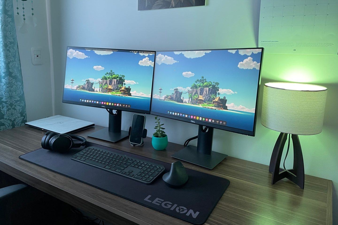 Mesa à meia luz, iluminada por um abajur à direita, com duas telas idênticas, teclado, mouse e headset sobre um mousepad grande.