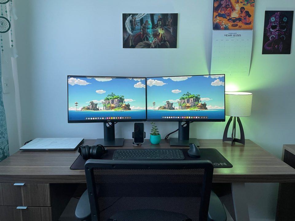 Outro ângulo do escritório do Matheus Fantinel, de frente, mostrando o pôster e o calendário na parede, acima dos monitores, e parte da cadeira.