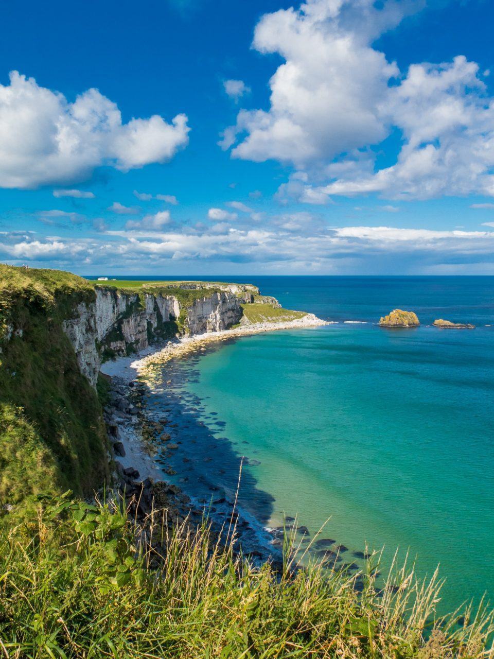 Foto tirada de cima de um penhasco, pegando todo o paredão do lado esquerdo da imagem, e uma praia de águas claras ao centro e à direita. Céu azul, com algumas nuvens brancas.