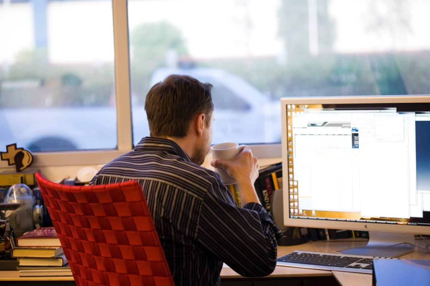 Elon Musk, de camisa listrada escura, sentado em uma cadeira vermelha, de costas para a câmera, encara a tela de um computador enquanto toma algo em uma xícara. Ao fundo, janelas amplas.