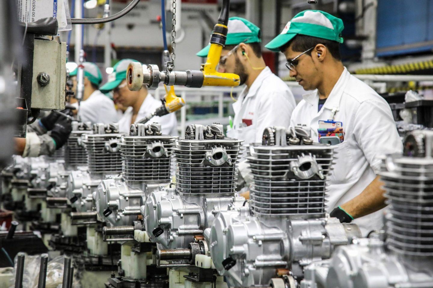 Foto de uma linha de montagem, com várias peças enfileiradas e alguns homens de macacões brancos e bonés verdes, mexendo nas peças.