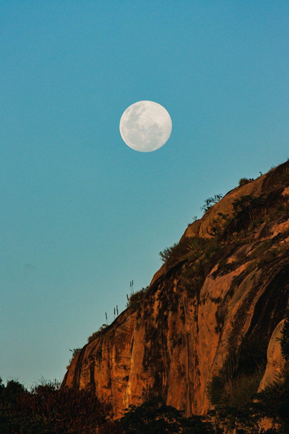 Rochedo ocupando a porção inferior direita da tela. Ao centro, um pouco acima, aparece a lua, branca e enorme, contra um céu azul não muito escuro.