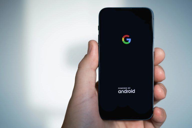 Mão segurando um iPhone 13 Mini, editado dificilmente para exibir o logo do Google e a inscrição “powered by Android”.
