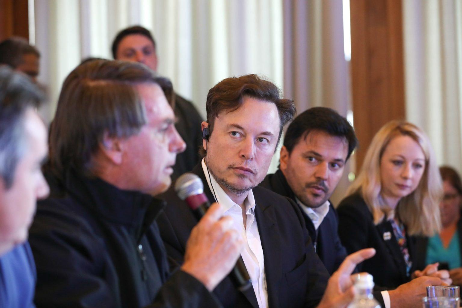 Foto lateral, com Elon Musk ao centro, focado, ladeado pelo presidente Jair Bolsonaro falando ao microfone e o ministro Fábio Faria.