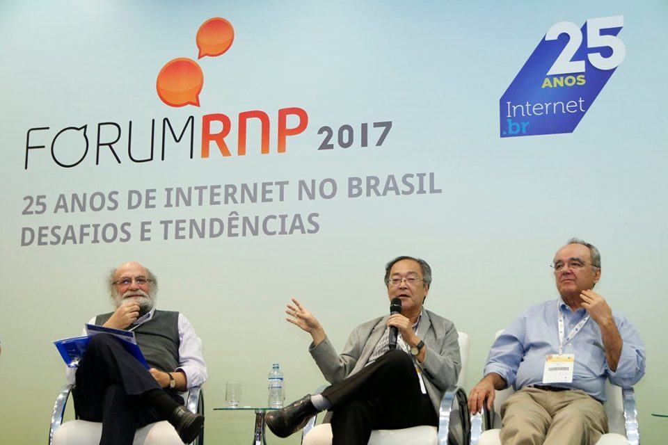 Demi, Tadao e Ivan sentados em cadeiras brancas. Ao fundo, ocupando a maior parte da imagem, um painel onde se lê “Fórum RNP 2017: 25 anos de internet no Brasil. Desafios e Tendências.”