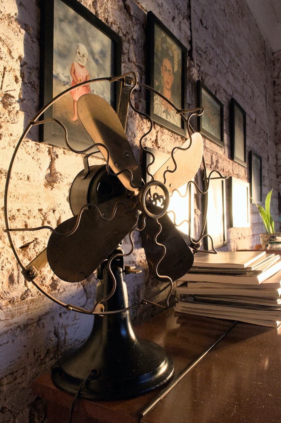 Ventilador antigo sobre um balcão, com algumas revistas e livros ao fundo e quadros pendurados numa parede com tijolos à mostra.