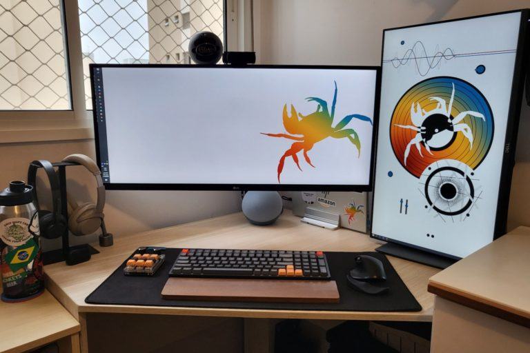 Mesa de trabalho de canto, vista de frente, com dois monitores grandes, um na horizontal e centralizado, outro à direita, na vertical. Teclado, mouse e um tecladinho menor à frente. Ao fundo, parede à direita e uma janela com tela de proteção à esquerda.