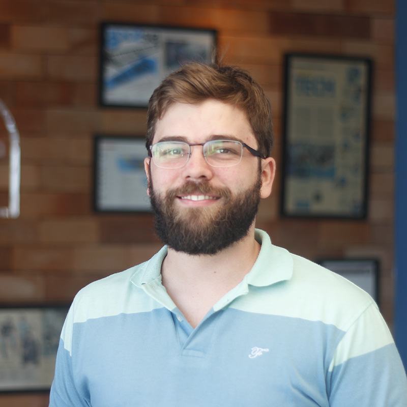 Homem branco, de óculos, bigode e barba e cabelo curto meio ruivo, vestindo uma camisa polo azul em dois tons. Ao fundo, parede de tijolinhos desfocada com alguns quadros pendurados.