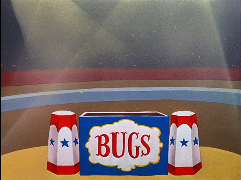 Picadeiro com caixas sob os holofotes. Na central, retangular, está escrito “Bugs” (referência ao Pernalonga).