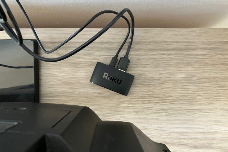 Visão de cima do Roku Express sobre um rack de madeira, atrás de uma TV, com cabos saindo de trás dele.