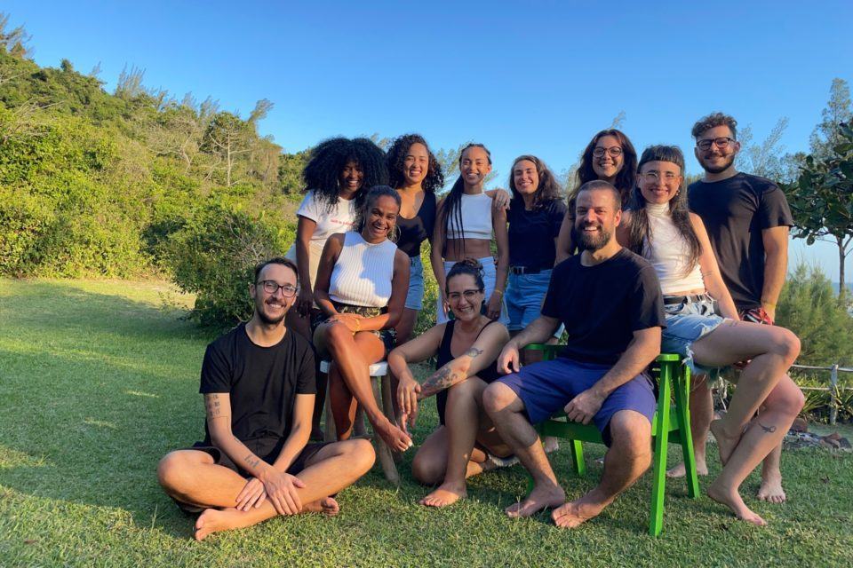 Grupo de pessoas sorridentes em um lugar a céu aberto, com muito verde e céu azul. Luciano está ao centro, à direita, sentado em uma cadeira verde.