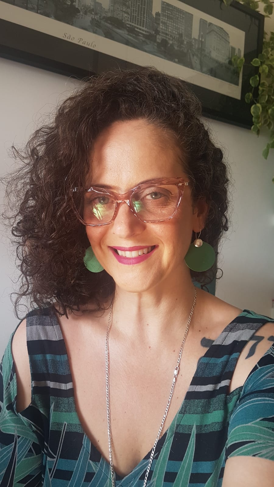 Mulher de óculos, cabelos encaracolados castanhos, vestindo uma roupa verde, com um colar discreto, sorrindo para a câmera.