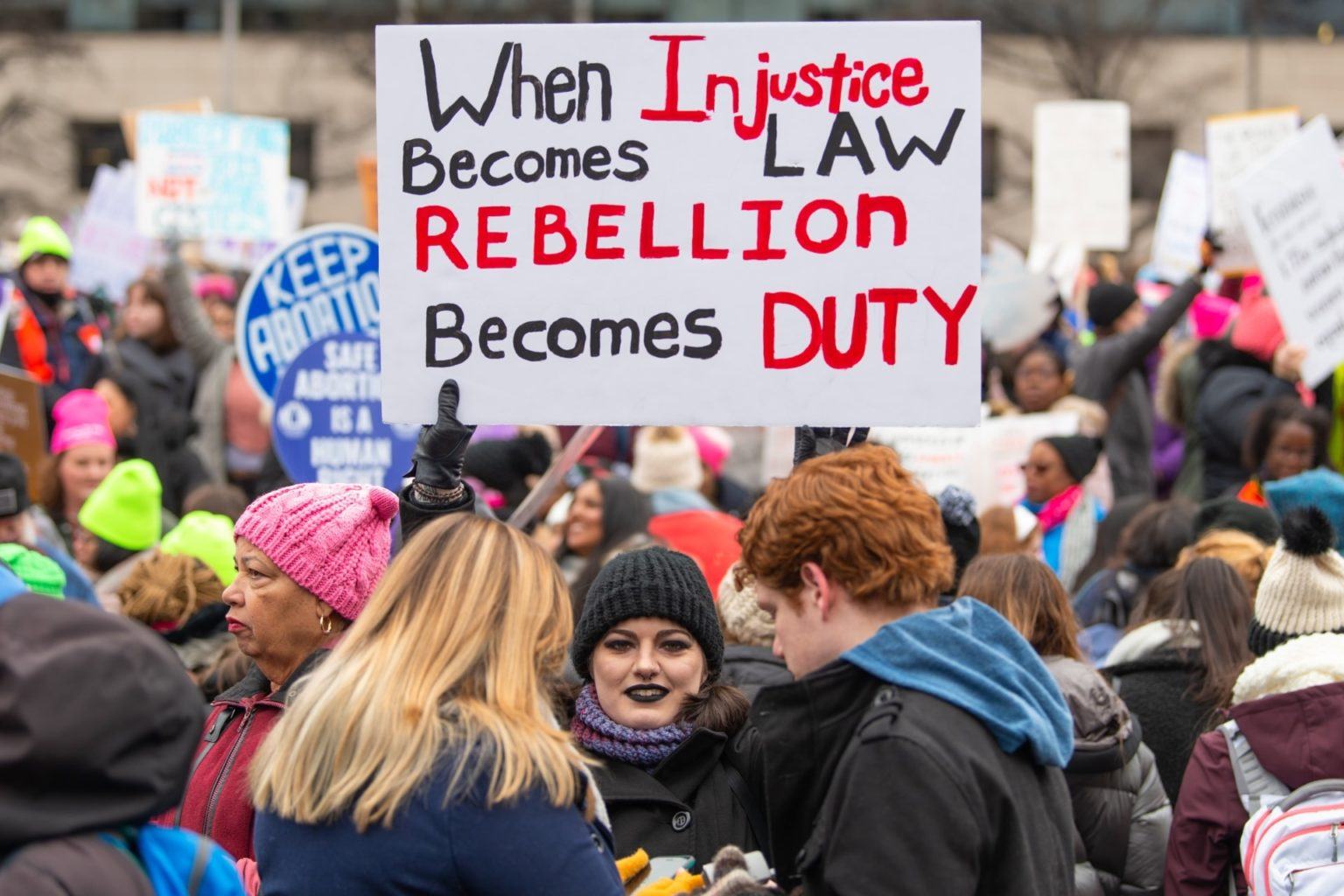Mulher branca, com batom e touca pretos e olhando para a câmera, segura um cartaz no meio da multidão. No cartaz, lê-se (em inglês): “Quando a injustiça vira lei, a revolta vira dever”.