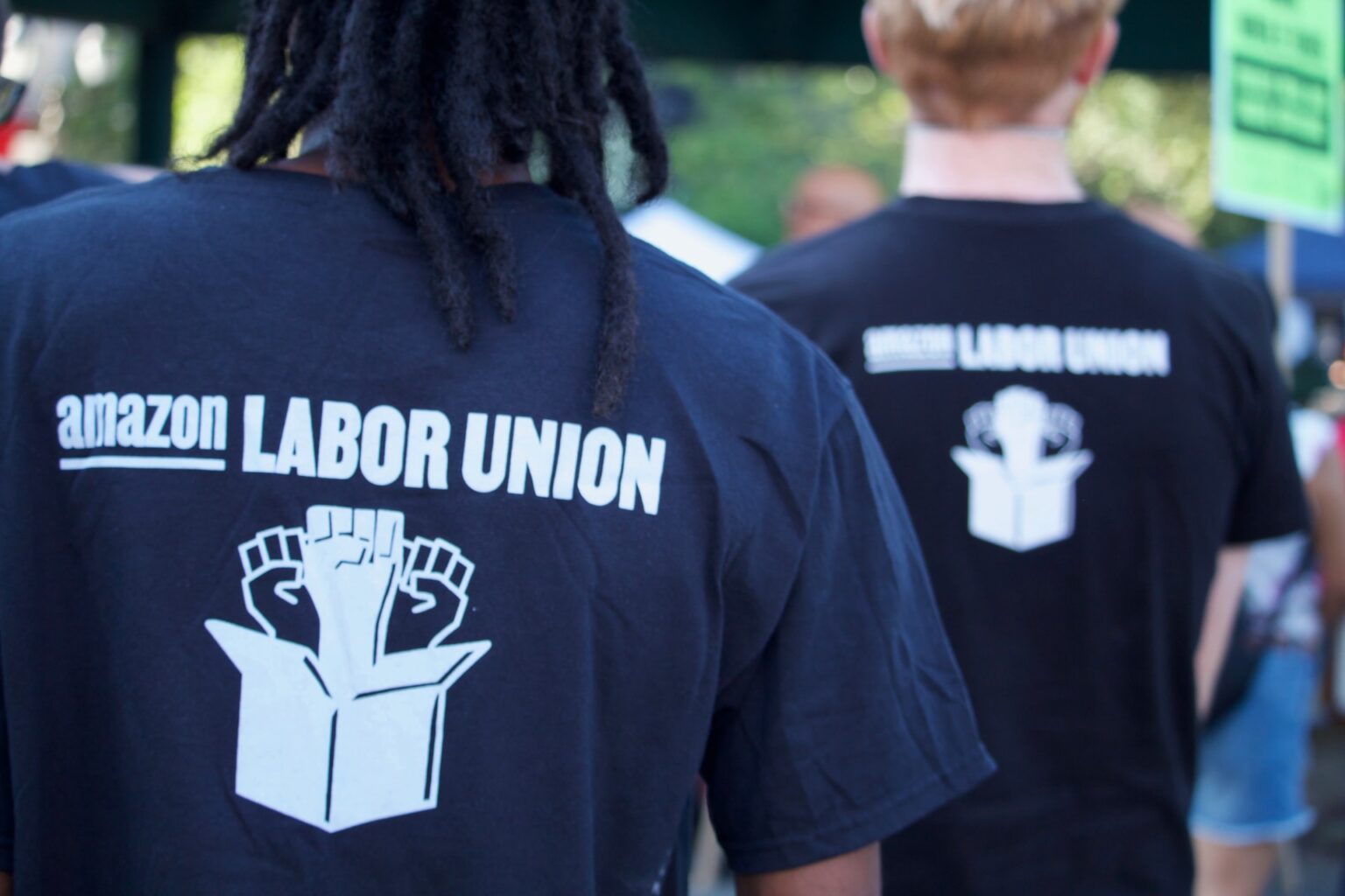 Dois homens de costas, vestindo camisetas com o logo do sindicato de funcionários da Amazon e o nome dele — “Amazon Labor Union”.