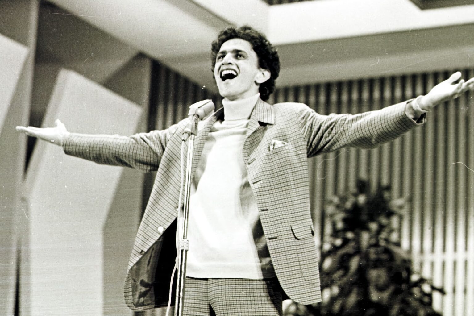 Um jovem Caetano Veloso, em foto em preto e branco, canta em um microfone com os braços abertos. Ele veste camiseta branca e um terno quadriculado claro.