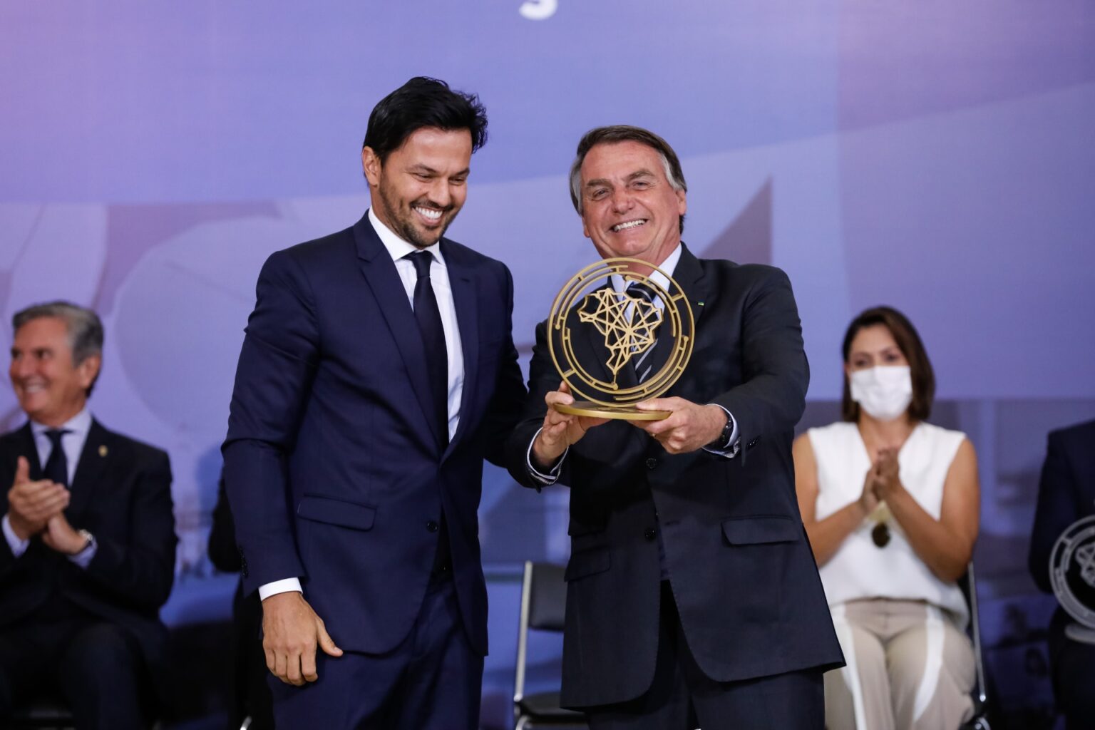 Dois homens brancos, de terno, rindo; o da direita (Jair Bolsonaro) segurando uma espécie de troféu e o da esquerda (Fabio Faria) olhando para baixo. Ao fundo, pessoas sentadas, desfocadas, batendo palmas.