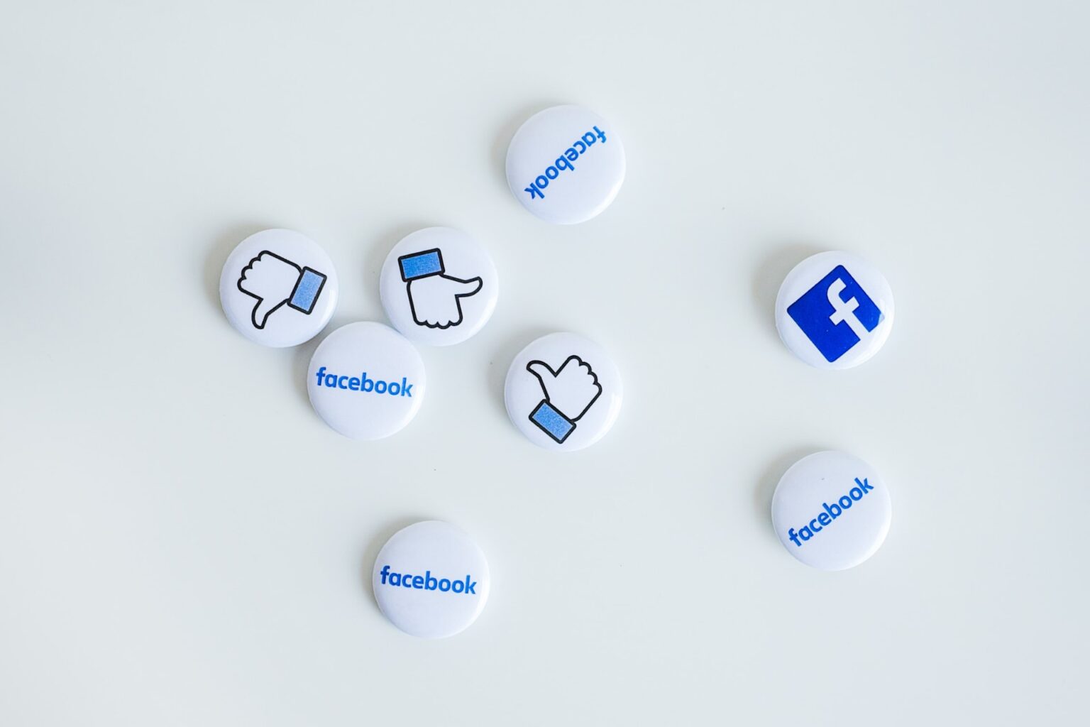 Alguns bottons do Facebook (com o logo, ícone de curtir e o nome da rede) espalhados em uma superfície branca.