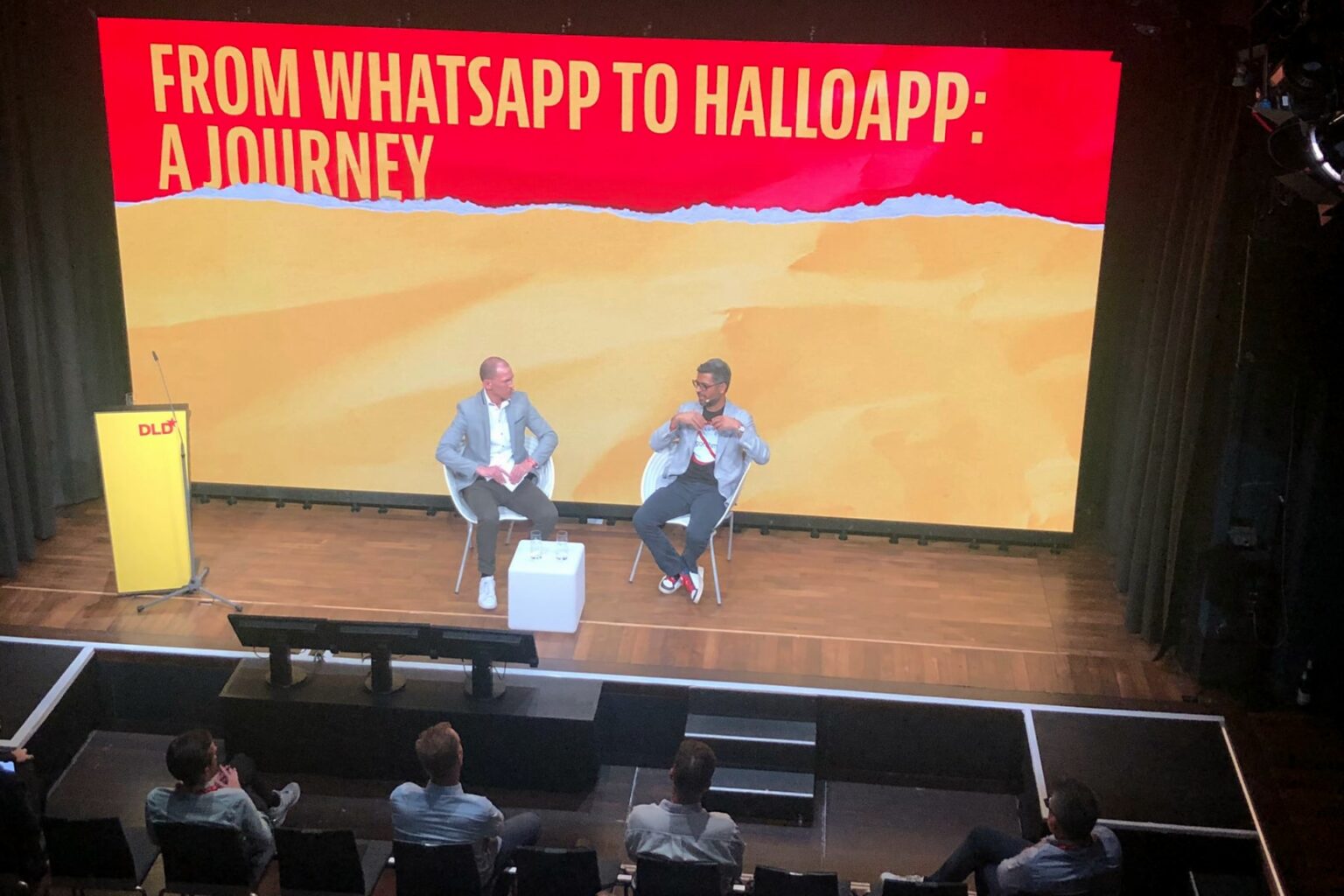 Foto de cima de um palco, com dois homens sentados no centro — o à direita da imagem é Neeraj. Ao fundo, uma tela enorme, amarela, com uma simulação de “rasgo” expondo um fundo vermelho e o título (em inglês) “Do WhatsApp ao HalloApp: Uma jornada”.