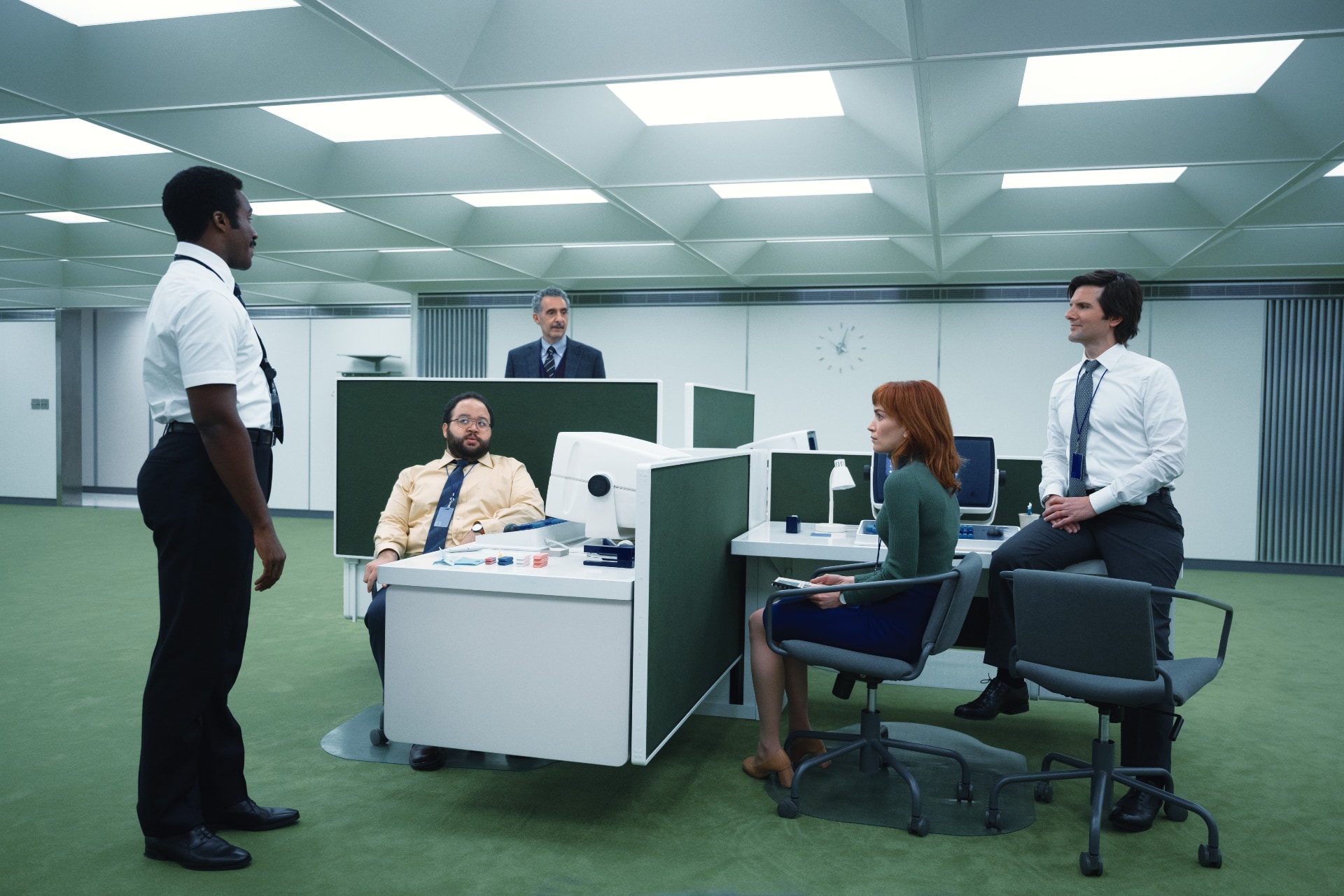 Four people (three men and one woman), in formal/office clothes, sitting or leaning on office tables separated by green partitions, in an old-fashioned office environment, green carpet and artificial lights on the ceiling. On the left, standing, a black man, also with formal clothes, supervises the group.
